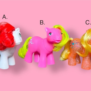 Puede incluir: Tres ponis de juguete coloridos sobre un fondo rosa. El poni A es blanco con pelo rojo, el poni B es rosa con pelo amarillo y el poni C es naranja con pelo amarillo. Cada poni tiene un diseño único.