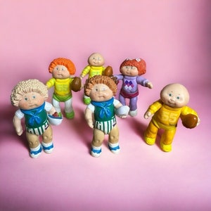 May include: Six Cabbage Patch Kids dolls dressed in sports uniforms, holding footballs and other sports equipment. The dolls are wearing various colors, including yellow, green, purple, and orange.