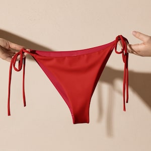 May include: A red bikini bottom with tie sides. The swimwear is held up by two hands against a neutral background. The bikini bottom is a solid red color with a smooth texture.