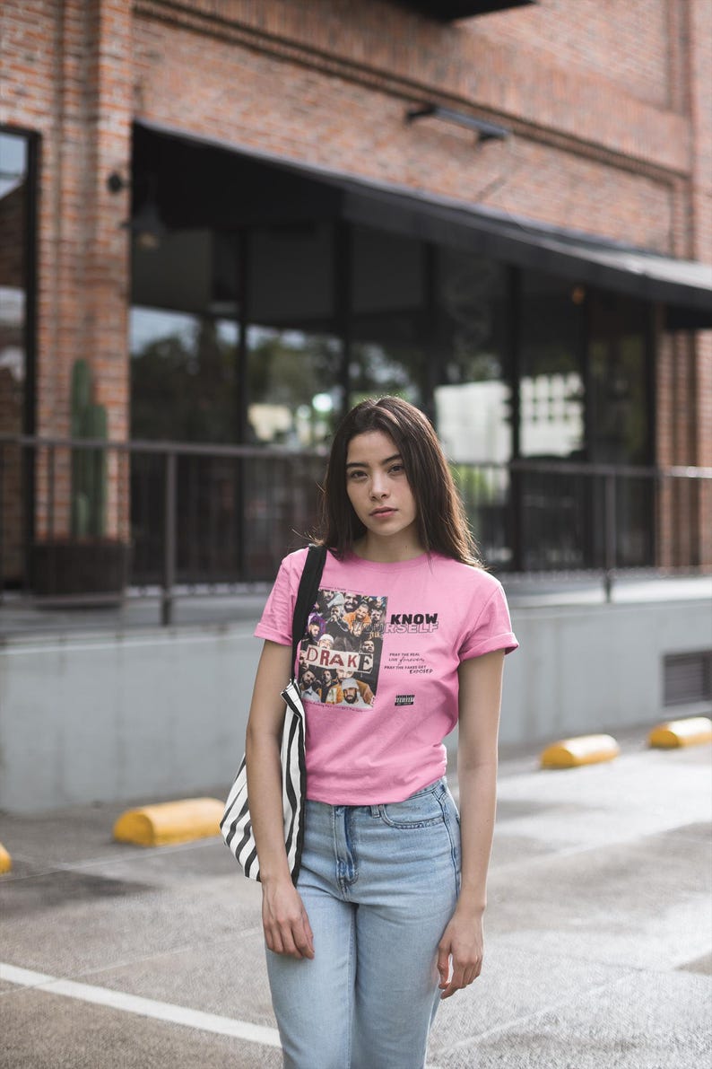 May include: A woman wearing a pink t-shirt with a graphic print of a group of people and the text "KNOW YOURSELF" and blue jeans. The graphic print includes the text "DRAKE".