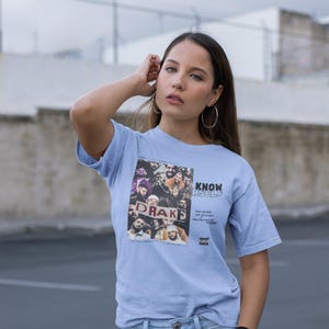 May include: A light blue t-shirt with a graphic print of a group of people holding a sign that says "Drake" and the text "Know Yourself" printed below the image. The text "Pray for the weak, Love your friends, Pray for the haters, Stay woke" is printed below the graphic.