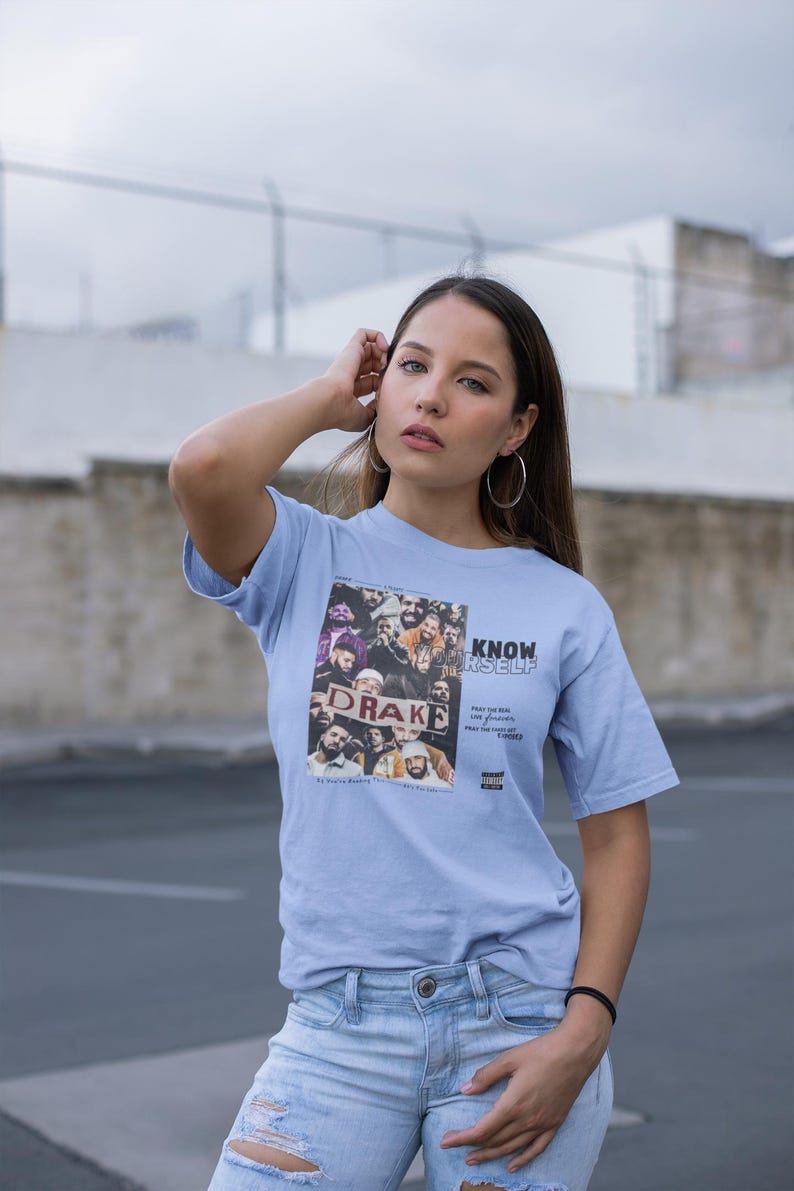 May include: A light blue t-shirt with a graphic print of a group of people holding a sign that says "Drake" and the text "Know Yourself" printed below the image. The text "Pray for the weak, Love your friends, Pray for the haters, Stay woke" is printed below the graphic.