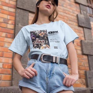May include: A light blue t-shirt with a graphic print of a group of people and the text "KNOW YOURSELF" and "DRAKE". The shirt is being worn by a person with brown hair and a black baseball cap. The person is wearing blue denim shorts and a black belt with a silver buckle.