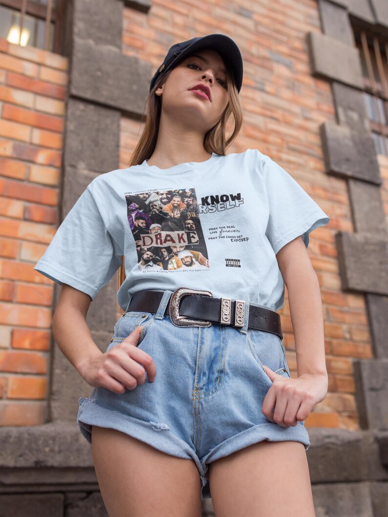 May include: A light blue t-shirt with a graphic print of a group of people and the text "KNOW YOURSELF" and "DRAKE". The shirt is being worn by a person with brown hair and a black baseball cap. The person is wearing blue denim shorts and a black belt with a silver buckle.