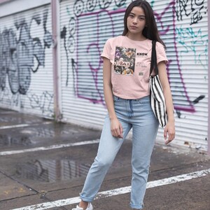 May include: A woman wearing a pink t-shirt with a graphic print of a group of people and the text "KNOW YOURSELF" and "DRAKE" in black. She is also wearing light blue jeans and white sneakers. She is standing in front of a graffiti-covered wall.