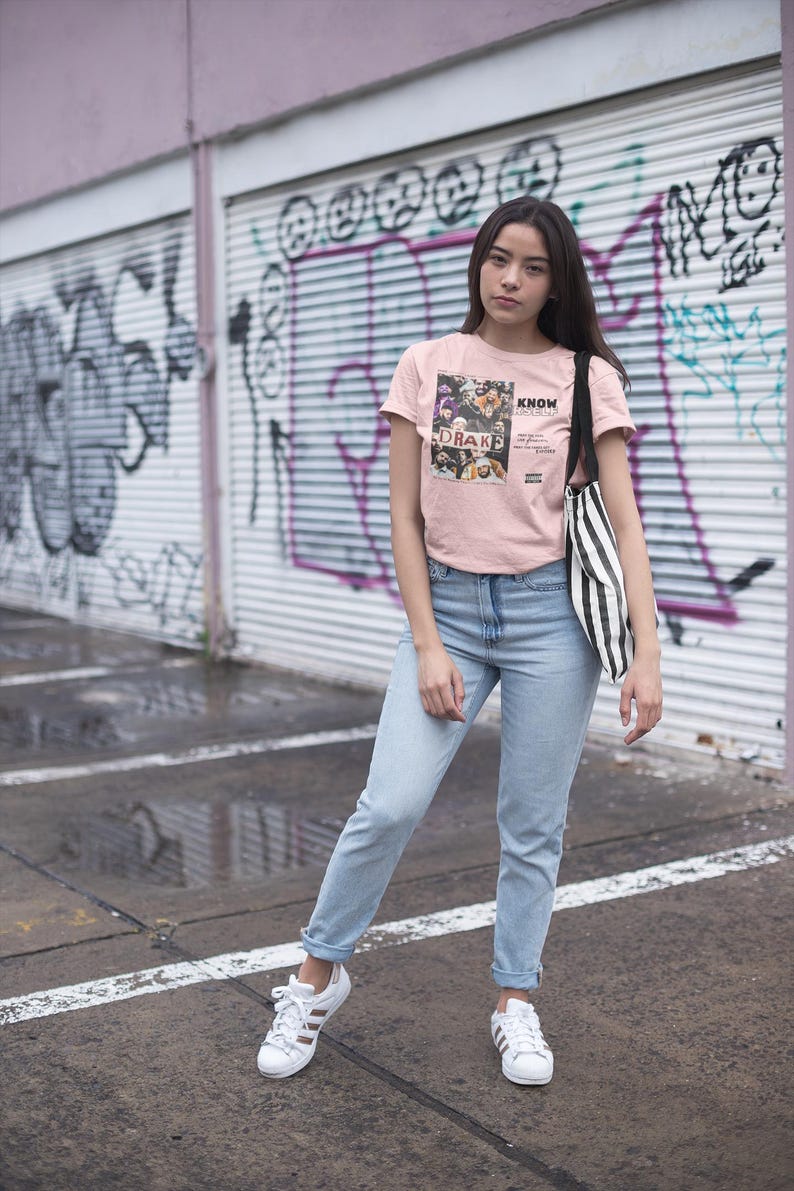 May include: A woman wearing a pink t-shirt with a graphic print of a group of people and the text "KNOW YOURSELF" and "DRAKE" in black. She is also wearing light blue jeans and white sneakers. She is standing in front of a graffiti-covered wall.