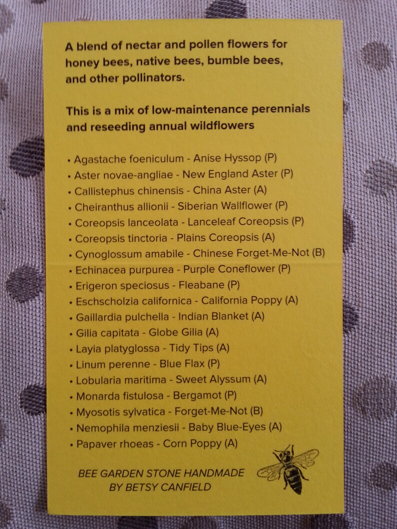 May include: A yellow card with a list of flowers and their common names, along with a letter code indicating their perennial (P) or annual (A) growth habit. The card also includes a small illustration of a bee. The text reads: "A blend of nectar and pollen flowers for honey bees, native bees, bumble bees, and other pollinators. This is a mix of low-maintenance perennials and reseeding annual wildflowers."  The card is titled "Bee Garden Stone Handmade by Betsy Canfield."