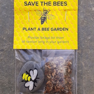 May include: A yellow and black bee illustration on a yellow honeycomb background with the text "SAVE THE BEES" and "PLANT A BEE GARDEN".  The text below the bee garden reads "Provide forage for them all season long in your garden!"  A small bag contains a grey painted rock with a bee illustration and a packet of wildflower seeds.
