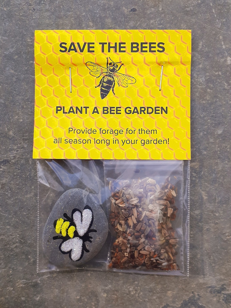 May include: A yellow and black bee illustration on a yellow honeycomb background with the text "SAVE THE BEES" and "PLANT A BEE GARDEN".  The text below the bee garden reads "Provide forage for them all season long in your garden!"  A small bag contains a grey painted rock with a bee illustration and a packet of wildflower seeds.