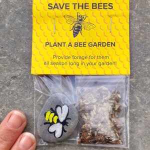 May include: A small yellow and black bee painted on a gray rock with a packet of wildflower seeds. The packet has a yellow background with the text "Save the Bees" and "Plant a Bee Garden" in black text. The text below reads "Provide forage for them all season long in your garden!"
