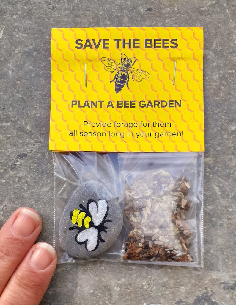 May include: A small yellow and black bee painted on a gray rock with a packet of wildflower seeds. The packet has a yellow background with the text "Save the Bees" and "Plant a Bee Garden" in black text. The text below reads "Provide forage for them all season long in your garden!"