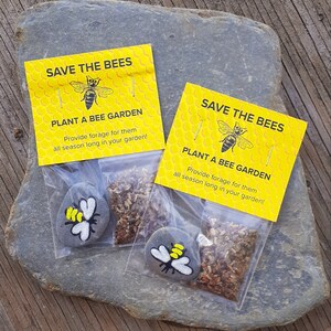 May include: Two small yellow and black bee-themed gift bags with the text "Save the Bees" and "Plant a Bee Garden" on the front. Each bag contains a small gray rock painted with a bee and a packet of wildflower seeds.
