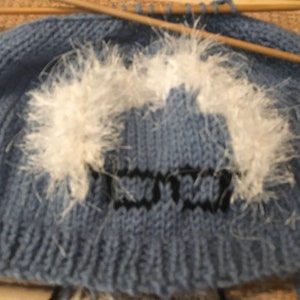 May include: A light blue knitted hat with a white fluffy design and black lettering. The hat is in the process of being knitted, with wooden knitting needles visible. The design appears to be a pair of glasses.