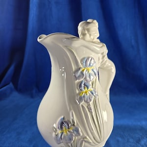 May include: A white ceramic pitcher with a sculpted woman figure on the handle and iris flower designs. The pitcher has a bulbous body and a curved spout. The irises are blue and yellow.