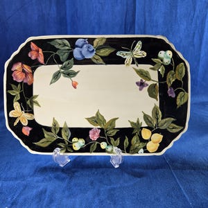 May include: Rectangular serving platter with a black border and a cream-colored center. The border is decorated with colorful flowers, butterflies, and leaves. The platter is supported by two clear plastic stands.