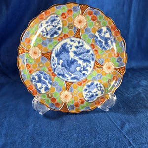 May include: A decorative ceramic plate with a scalloped edge. The plate features a central blue and white floral design, surrounded by smaller floral and geometric patterns in orange, green, and blue. The rim has a gold border.