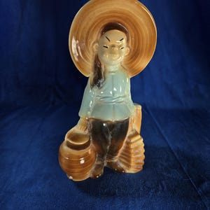 May include: A ceramic figurine of a person wearing a brown hat and blue clothing, holding a brown pot and a brown fan. The figurine is a planter.