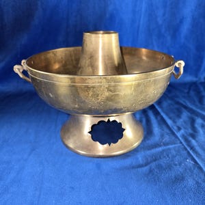 May include: A brass hot pot with a central chimney and two handles. The pot sits on a round base with an openwork design.