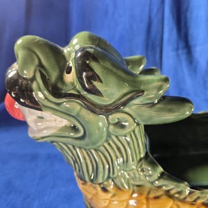 May include: A ceramic dragon-shaped planter in shades of green, yellow, and red. The dragon's head is green with black accents and a red nose. The body is yellow with a scale pattern. The planter is designed to hold plants or other decorative items.