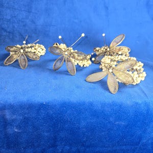 May include: Four silver butterfly napkin rings with pearl accents. The rings are on a blue background.