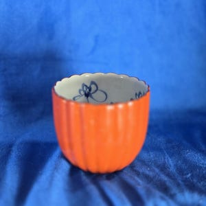 May include: A small orange ceramic cup with a white interior and blue floral design. The cup has a fluted exterior.