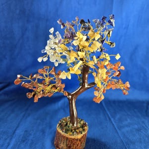 May include: A decorative gemstone tree with a brown trunk and branches, adorned with colorful gemstone leaves in shades of clear, yellow, orange, purple, and blue. The tree sits in a small, brown, bark-like base, creating a miniature, naturalistic sculpture.