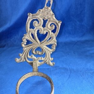 May include: Ornate, cast iron wall-mounted candle holder with a decorative, scrolled design. The holder features a circular ring for the candle and a detailed, heart-shaped embellishment. The metal has a distressed, aged appearance.
