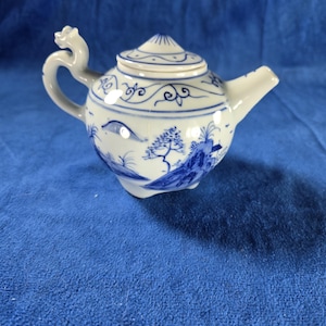 May include: A small, white porcelain teapot with blue painted landscape and floral designs. The teapot has a curved spout, a dragon-shaped handle, and a lid with a small knob. The design is reminiscent of traditional Asian pottery.