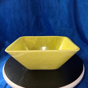 May include: A square, green ceramic bowl with a speckled glaze. The bowl is sitting on a black and white turntable.