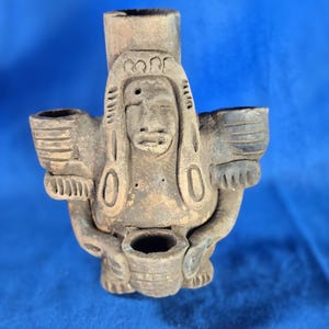 May include: A weathered, brown ceramic figurine with a stylized human face and headdress. The figure has two side compartments and a central cylindrical element. The piece appears to be handmade and has a rustic, aged appearance.