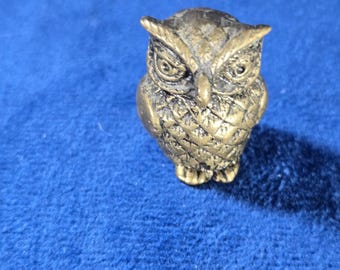 May include: A small, bronze-colored owl figurine. The owl has detailed feather patterns and a stern expression. The figurine is standing on a blue surface. The owl is a decorative item.