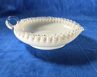 May include: A white, oval-shaped serving dish with a clear handle. The dish has a ruffled edge and a spiral design in the center. The dish is sitting on a blue surface.