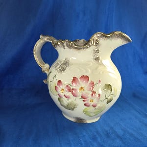 May include: White ceramic pitcher with a gold rim and floral design. The pitcher has a handle and a spout. The flowers are pink and white with green leaves.