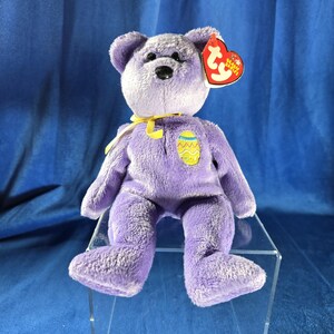 May include: Purple Beanie Baby bear with a yellow ribbon and an Easter egg design. The bear has a black nose and black eyes. The tag says "Ty Beanie Babies".