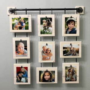 Könnte beinhalten: Eine Wandmontage mit neun weißen Holzrahmen, die an einem schwarzen Metallrohr hängen. Jeder Rahmen hat einen schwarzen Clip, um ein Foto zu halten. Die Fotos zeigen eine Familie.