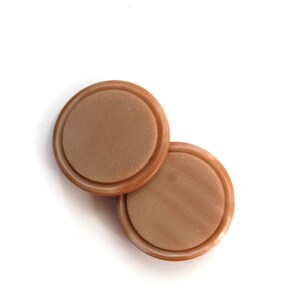 May include: Two round, light brown buttons with a raised outer rim and a flat, textured center. The buttons are slightly overlapping against a white background, showcasing their simple, classic design.