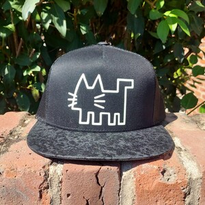 May include: Black trucker hat with a white cat graphic. The hat has a black mesh back and a patterned brim. The cat design is a simple, geometric outline. The hat is sitting on a brick wall with green foliage in the background.