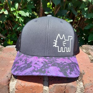 May include: A gray trucker hat with a black mesh back and a purple and black patterned brim. The hat features a white graphic of a cat. The hat is resting on a brick surface.