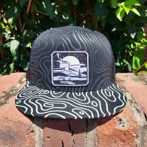 May include: Black trucker hat with a white and black topographic pattern on the brim and a matching pattern on the crown. The hat features a square patch with a landscape design in white and gray.