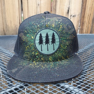 May include: Black and green camouflage trucker hat with a green circle patch featuring three black pine trees. The hat has yellow paint splatters.