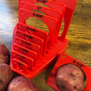 May include: Red plastic potato sizing guide with nine different sized rings. The rings are labeled with measurements in inches, including 3/4 inch, 1-1/2 inch, 1-5/8 inch, 1-3/4 inch, 1-7/8 inch, 2-1/4 inch, 2-3/4 inch, 3 inch, 3-1/4 inch, and 4-1/2 inch. There are three red potatoes next to the sizing guide.
