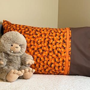 May include: A brown and orange pillow with a pumpkin pattern and a brown border. A gray plush toy is sitting on the pillow.