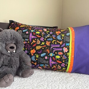 Peut inclure: Un jouet en peluche de loup gris est assis sur un lit avec un oreiller noir et violet avec un motif de sorcière coloré. L'oreiller a une bordure verte, orange et jaune.