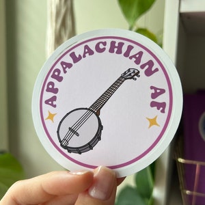 May include: A white round sticker with a pink border and the text "Appalachian AF" in a retro font. A banjo is illustrated in the center of the sticker.