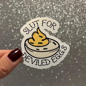 May include: A white sticker with a black outline and the text "Slut for Deviled Eggs" in a vintage tattoo style font. A cartoon deviled egg with yellow yolk and green chives is depicted in the center.