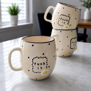 May include: Cream-colored ceramic mugs with black polka dots and the phrase "fuck it" in a dotted frame. Each mug has a small daisy accent. The mugs are stacked on a marble surface.