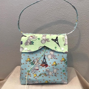 May include: A light blue shoulder bag with a green flap. The bag is decorated with a pattern of white poodles, Eiffel Towers, and gold coins. The flap has a pattern of white poodles and green balls of yarn.