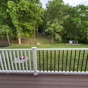Puede incluir: Vista de un jardín desde una terraza con un trampolín, árboles y una pequeña estructura. La terraza tiene barandillas blancas y negras, y el jardín está cubierto de hierba verde.