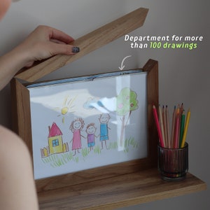 May include: A wooden picture frame with a shelf underneath. The frame is open and a drawing of a family is visible. A glass jar filled with colored pencils sits on the shelf. The text "Department for more than 100 drawings" is written on the frame.