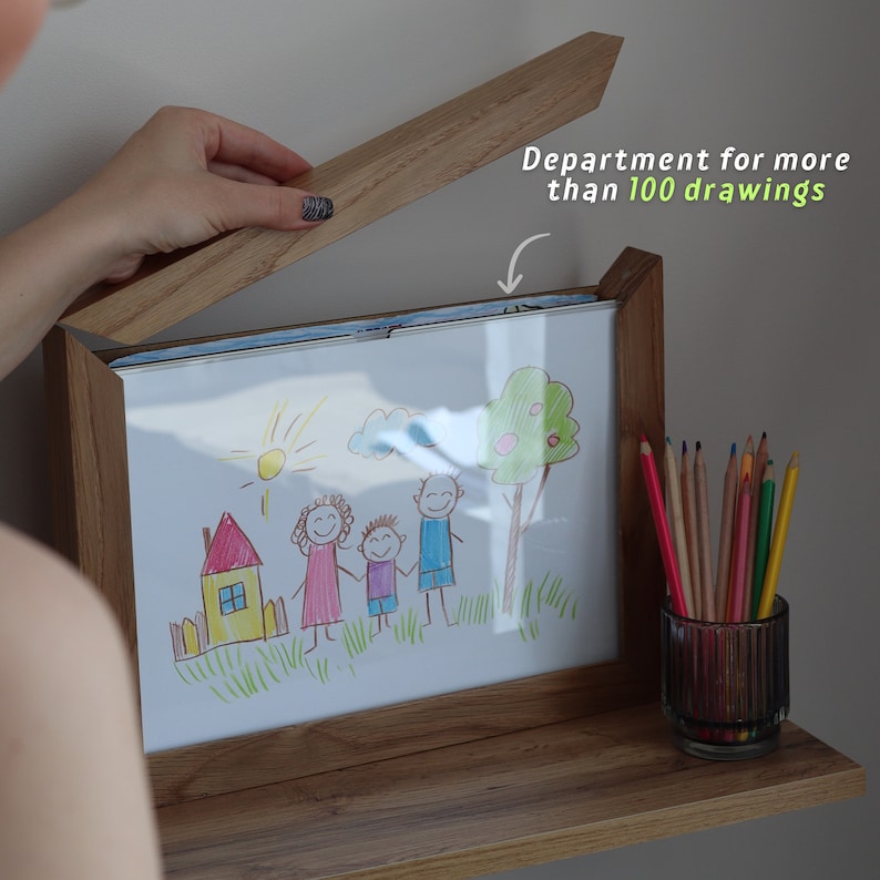 May include: A wooden picture frame with a shelf underneath. The frame is open and a drawing of a family is visible. A glass jar filled with colored pencils sits on the shelf. The text "Department for more than 100 drawings" is written on the frame.